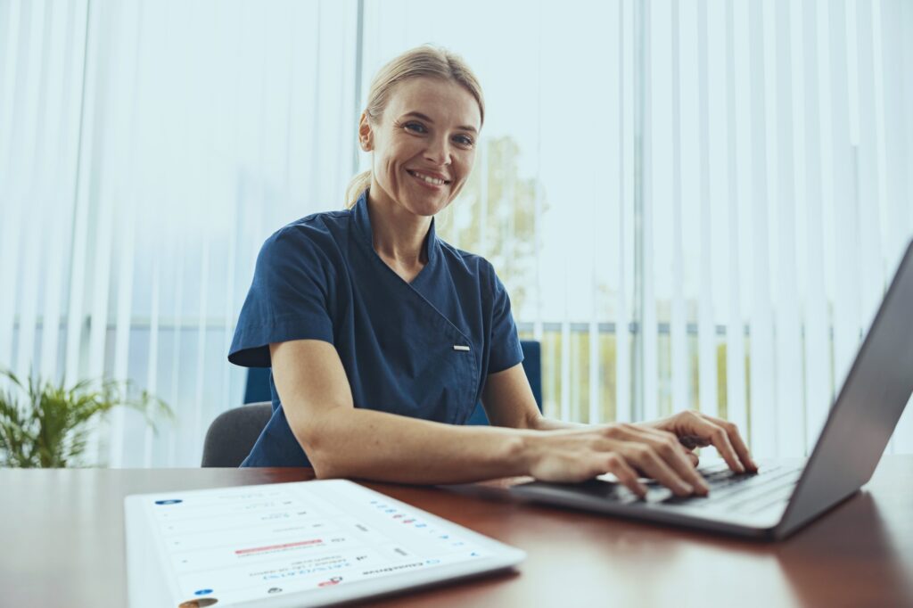 Smiling doctor therapist working laptop during appointment in her medical office and looking camera