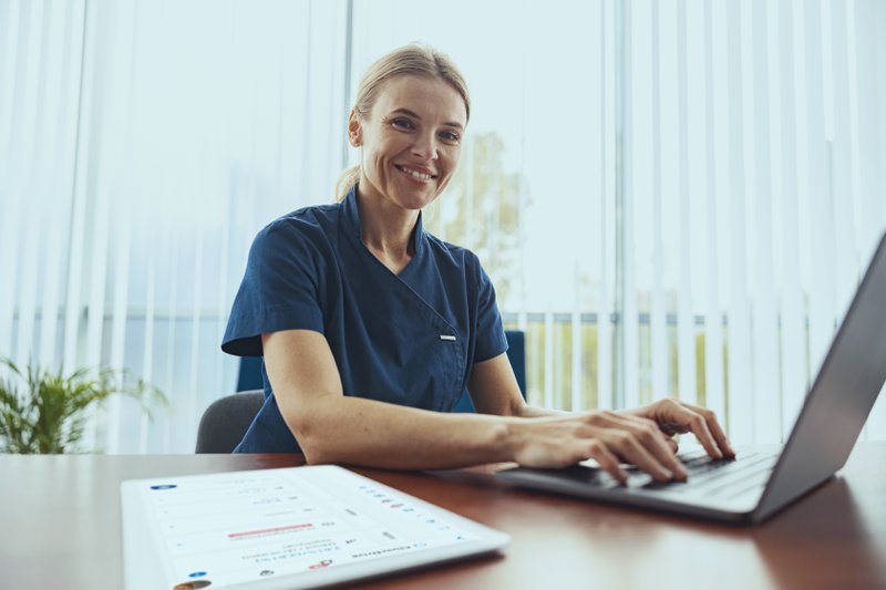 Smiling doctor therapist working laptop during appointment in her medical office and looking camera